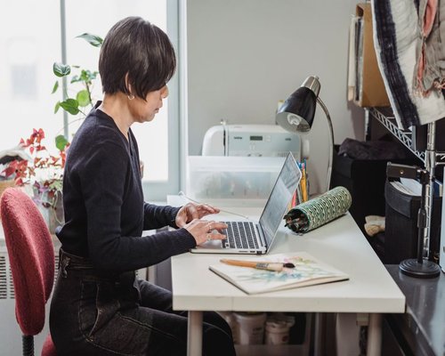A person sitting with good ergonomic posture at an office desk using a laptop with natural light coming from the side